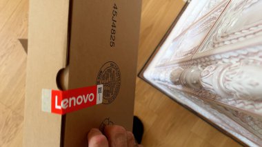 Paris, Fransa - 4 Nisan 2025: Mühürlü bir Lenovo karton kutuyu tutan el, görünür kırmızı Lenovo damgalı, dekoratif seramik bir soba ve ahşap bir zeminin yanına yerleştirilmiş.