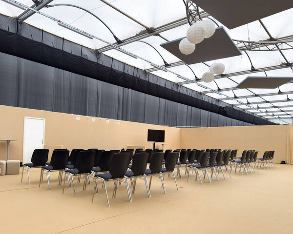 Современное закрытое пространство для мероприятий с рядами черных стульев, стоящих перед экраном монитора, под прозрачной крышей с подвесными акустическими панелями и бумажными фонарями в нейтральной обстановке