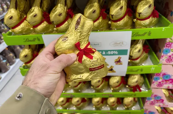 Paris, Fransa - 28 Mart 2025: Bir el Lindt Lapin 'i ya da Fransız süpermarketinin Paskalya temalı şeker bölümünde kırmızı kurdeleli altın folyoya sarılı çikolatalı tavşanı tutar