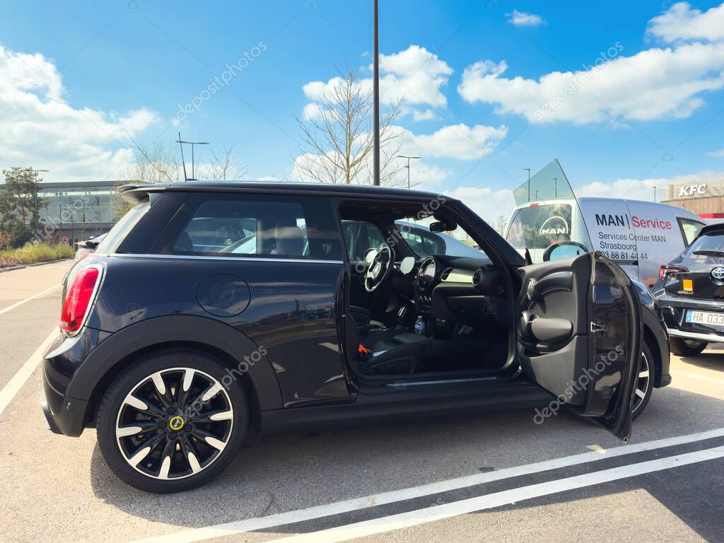 Paris, France- Mar 27, 2025: A black electric Mini Cooper parked outdoors with the driver door open in a shopping zone near a service van and dealership under a sunny blue sky