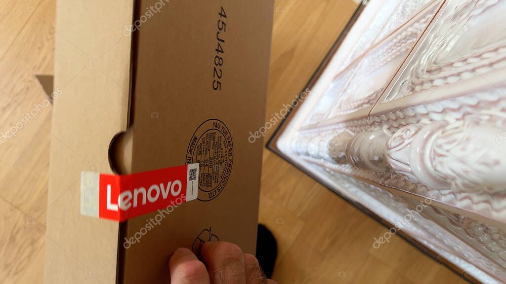 Paris, France - Apr 4, 2025: A close-up view of a hand holding a sealed Lenovo cardboard box with visible red Lenovo branding, placed next to a decorative ceramic stove and a wooden floor