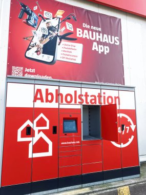 Berling, Almanya - 5 Nisan 2025: Dokunmatik ekran ve açık dolap kapısı olan Kırmızı Bauhaus pikap istasyonu, Almanya 'da ürün taraması ve çevrimiçi alışveriş için Bauhaus uygulamasını tanıtan büyük bir reklamın altında duruyor.