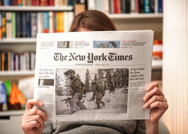 Paris, Fransa - 22 Nisan 2025: Bir kadın içeride oturmuş The New York Times 'ın uluslararası basımını okuyor, arka planda Vitsoe kitaplıklarının bulunduğu bir ticaret savaşı ve İran nükleer müzakereleri ile ilgili ön sayfa makaleleri yer alıyor.