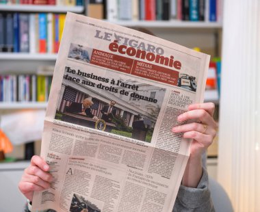 Paris, Fransa - 22 Nisan 2025: Le Figaro gazetesinin ekonomi bölümünde ABD gümrük vergileri ve iş aksamaları ile ilgili ön sayfada yer alan ve arka planda kitap raflarının görülebildiği bir makale yayınlanan bir kişi,