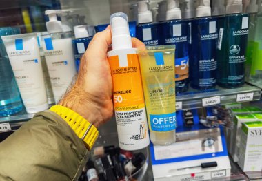 Paris, Fransa - 28 Kasım 2023: Bir eczanede iki La Roche-Posay cilt ürününü tutan sarı saat takan bir el, Anthelios SPF 50 artı sprey ve bir Lipikar 'ın diğer La Roche-Posay ürünleriyle dolu bir rafın önünde yağ şişesini temizlemesi.