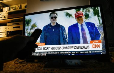 Paris, Fransa - 19 Şubat 2025: Bir adam dijital olarak değiştirilmiş bir CNN haber yayınını izlerken SpaceX tişörtlü Elon Musk ve kırmızı MAGA şapkalı Donald Trump 'ın montajlanmış görüntülerini seyrediyor.