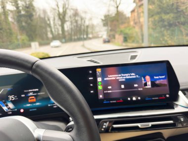 Elektrikli bir arabanın içinde, direksiyon ve sürücü ekran çalışırken odaklanmış haber içeriğini gösteren büyük bir merkezi ekran görüntüsü var.