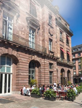 Strasbourg, Fransa - 4 Nisan 2025: Strasbourg 'daki eski Bourse binasının güneşli manzarası şimdi Brasserie Louise' de insanların açık mavi bir gökyüzü altında kalabalık bir açık hava terasında öğle yemeğinin keyfini çıkarıyorlar.
