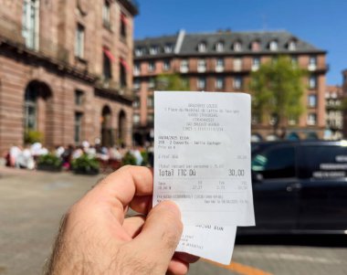 Strasbourg, Fransa - 4 Nisan 2025: Strasbourg 'daki Brasserie Louise' den iki öğün yemek fişi tutan erkek, tarihi binalar ve açık hava lokantalarını gün ışığında gösteren bulanık bir geçmişe sahip.