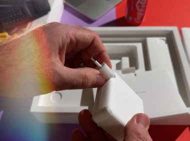 Paris, Fransa - 4 Nisan 2025: Coca-Cola kutusu olan renkli bir çalışma alanındaki boks oturumunda koruyucu kağıda sarılı beyaz bir MacBook şarj aletine iki el dikkatlice Avrupa fişi takıyor..