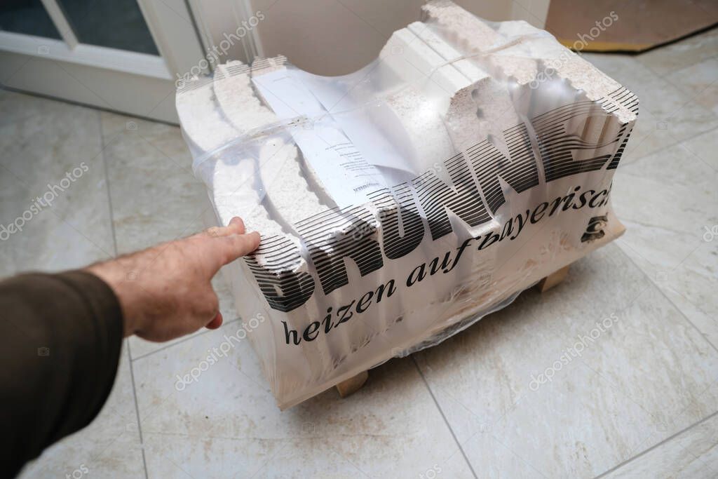 Munich, Germany - Apr 25, 2023: Hand pointing towards a stack of Brunner fireclay bricks wrapped in plastic with the slogan Heizen auf bayerisch visible, placed on a wooden pallet on a tiled floor.