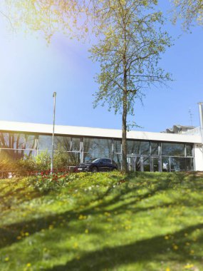 Berlin, Almanya - 12 Nisan 2025: Modern cam önlü bir binanın önüne park edilmiş elektrikli Mercedes-Benz SUV, kır çiçekleriyle kaplı canlı yeşil bir yamaç ve açık mavi gökyüzünün altındaki ağaçların gölgeleri.
