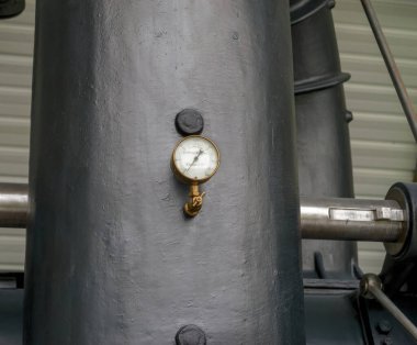 Paris, Fransa - 20 Nisan 2025: Arkaplanda çelik pistonlar ve tırtıklı bir duvar bulunan kalın siyah metal bir sütuna monte edilmiş pirinç basınç göstergesi gösteren endüstriyel bir buhar makinesinin yakın çekimi