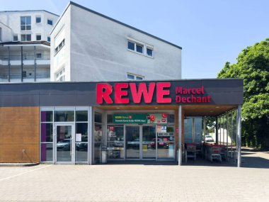 Rastatt, Almanya - Mayıs 12025: Rastatt 'taki REWE Marcel Dechant süpermarketinin ön cephesinde güneşli bir günde kırmızı tabela, cam giriş, alışveriş arabaları ve bitişik konut araçları yer alıyor