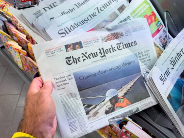 Berlin, Almanya - 16 Mayıs 2025: 16 Mayıs 2025 tarihli New York Times International Edition 'ı elinde tutan bir kişi, bir güneş enerjisi hikayesi nükleer tehdit görüşünü ve Suriye ile ilgili Trump diplomasisini dergi rafında gösteriyor