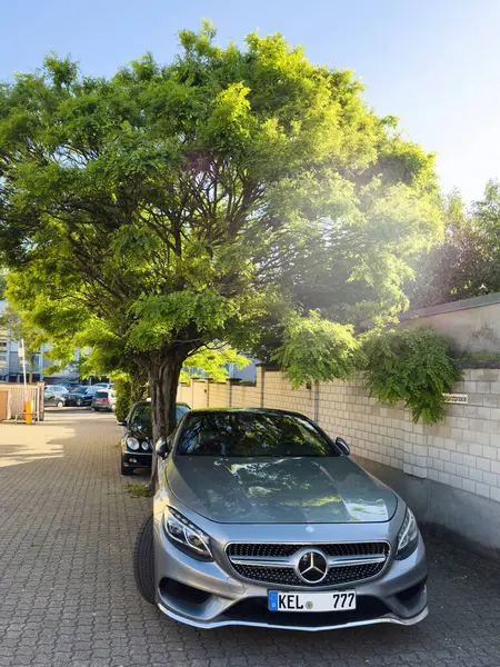 Kehl, Almanya - 11 Mayıs 2025: Zarnarztpraxis etiketli beyaz tuğlalı bir duvarın yanında, sık bir ağaç tepe örtüsünün altındaki gölgeli bir sokakta parlak bir gümüş Mercedes-Benz park edilmiştir.