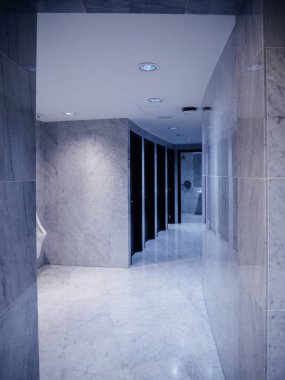 Halka açık bir tuvaletin mermer döşeli koridorunda parlak siyah kabin kapıları tavan lambaları ve beyaz pisuarın yanından geçen yansıtıcı duvarlar var.