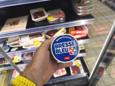 Paris, Fransa - 26 Mayıs 2025: Sarı saat takan bir el, çeşitli paketlenmiş et ve gıda ürünleriyle dolu soğutulmuş bir süpermarketin içinde 300 gram Bresse Bleu peyniri bulundurur.