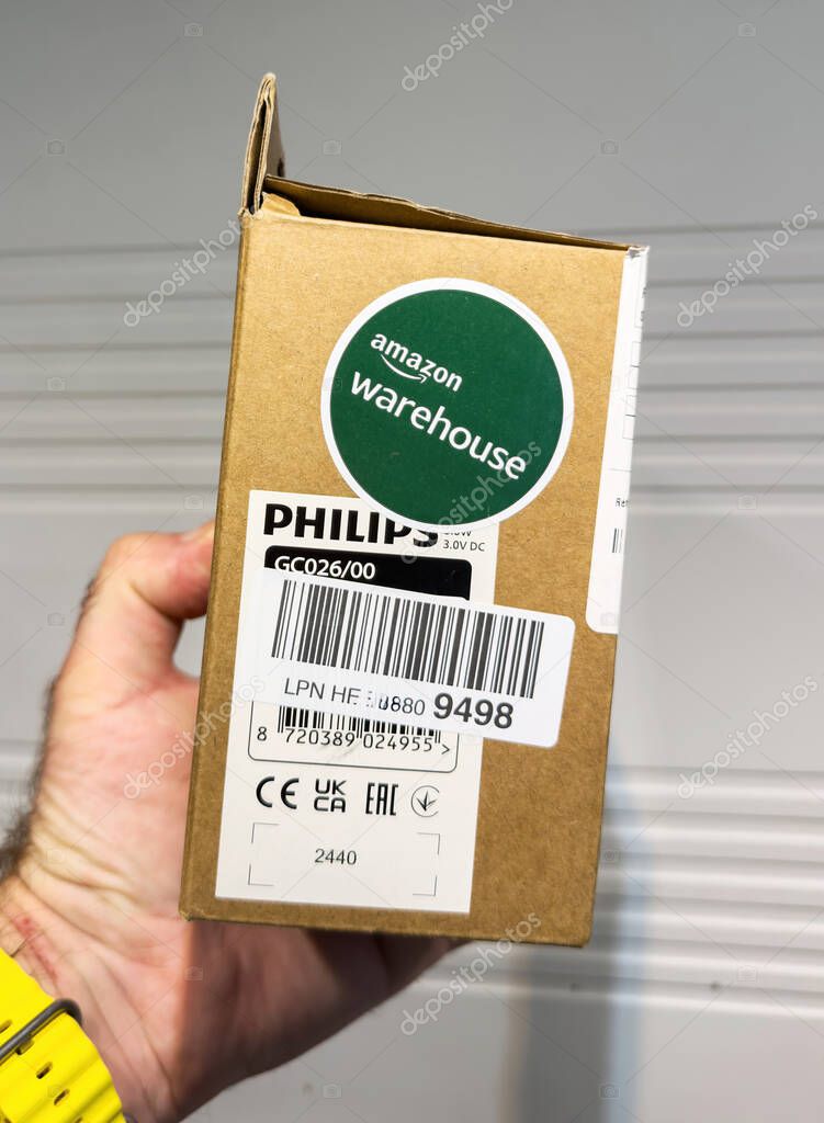 Ulm, Germany - May 6, 2025: A hand holds a Philips GC026 box with Amazon Warehouse sticker, barcode labels, and certification symbols, set against a softly lit, neutral background with horizontal lines visible