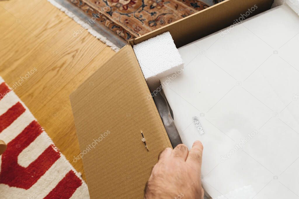 Paris, France - Nov 29, 2024: Person opening a cardboard box containing a Canton subwoofer wrapped in white protective fabric, with foam corner protectors, on a wooden floor near patterned and red carpets