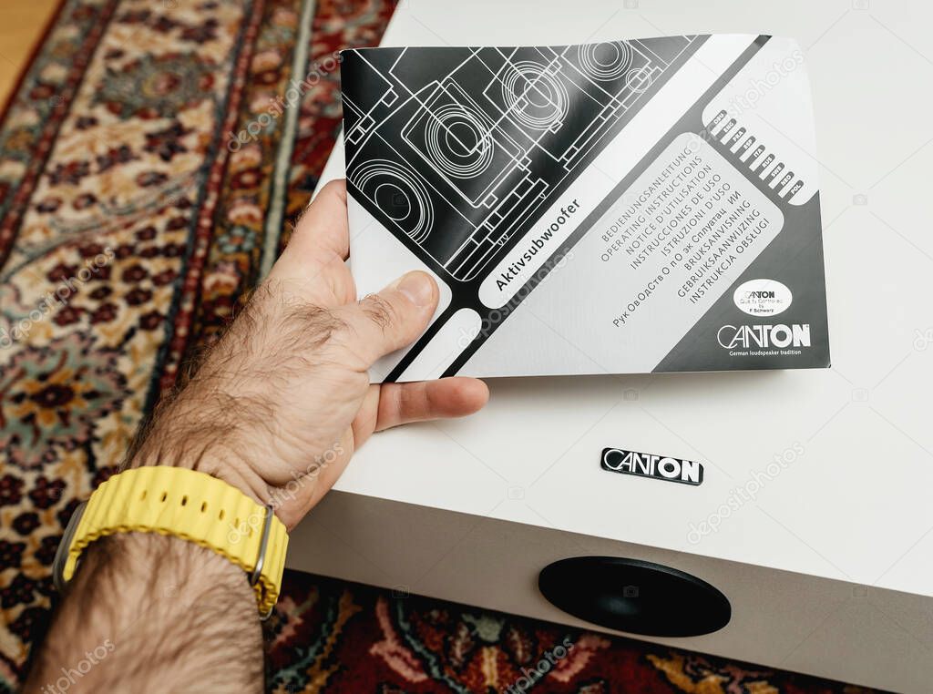 Paris, France - Nov 29, 2024: Person with a yellow watch holding the instruction manual for a Canton active subwoofer, with the white subwoofer and Canton logo visible on a patterned rug background.