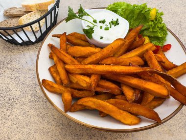 Bir tabak çıtır patates kızartması kremalı sosla servis edilir ve marul ve otlarla süslenir, yanında da taş bir masanın üzerinde çeşitli ekmek dilimleri bulunur..