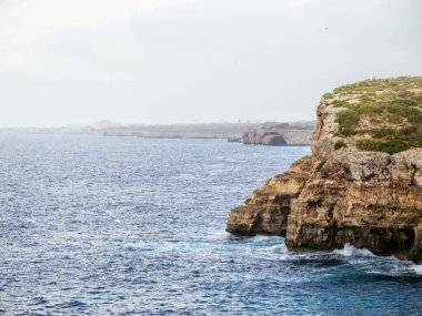 Sarp kayalıkları olan dramatik bir kayalık kıyı şeridi derin mavi dalgalara bakar. Uzak bir kentsel ufuk çizgisi ise Mallorca 'da sisli bir gökyüzünün altında belli belirsiz görünür.