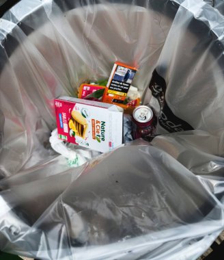 Berlin, Almanya - 18 Nisan 2025: Açılmış gıda ambalajı, bir soda kutusu, içinde sağlık uyarısı olan bir sigara paketi ve diğer görünür atıkların bulunduğu açık plastik bir çöp tenekesi.