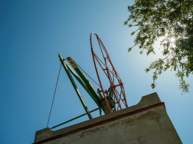 Kırmızı ve yeşil bileşenlere sahip geleneksel bir metal yel değirmeni yapısı, İspanya 'nın Mallorca kentindeki açık mavi gökyüzüne karşı yükseliyor ve güneş ışığı ağaç dallarının arasından süzülüyor.