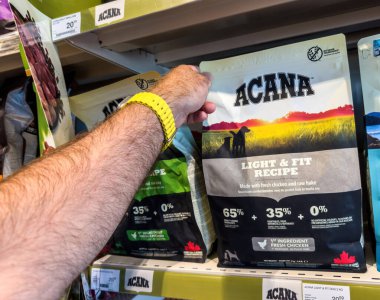 Manacor, İspanya - 17 Temmuz 2025: Elinde Acana Light and Fit köpek maması çantası ve taze tavuk ve ham hake malzemeleri olan bir kişinin hayvan mağazasının rafında görüntülenmesi