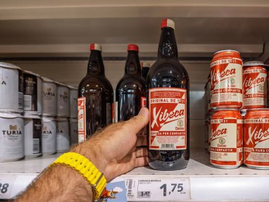 Inca, İspanya - 21 Temmuz 2025: İspanya 'nın bir süpermarketinde yanında eşleşen teneke kutular ve diğer bira markaları bulunan büyük bir Xibeca bira şişesini tutmak