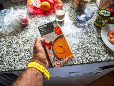 Muro, İspanya - 24 Temmuz 2025: Bir el, İspanyol mutfağında Eroski 'den alınmış bir deniz ürünleri karton çorbasını granit tezgahta pişirme malzemeleri ve çatal bıçaklarıyla tutuyor.