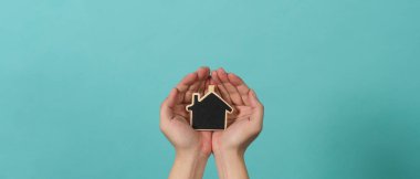 Small wood house in hands represent concepts such as home care family love real estate housing shelter insurance and mortgage. Hands holding small model house isolated on blue green studio background.