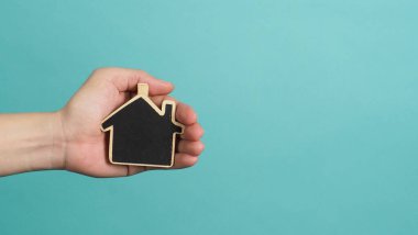 Small wood house in hands represent concepts such as home care family love real estate housing shelter insurance and mortgage. Hands holding small model house isolated on blue green studio background.