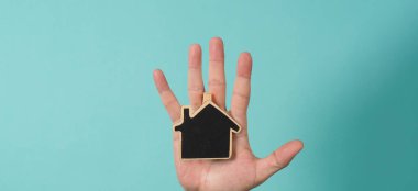 Small wood house in hands represent concepts such as home care family love real estate housing shelter insurance and mortgage. Hands holding small model house isolated on blue green studio background.