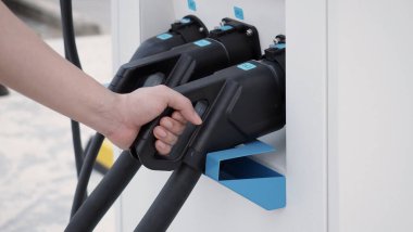 El ele tutuşan EV şarj prizi dışarıda bulanık bir arka plan ile kameranın önünde. Elektrikli araba sürücüsünde şarj prizi vardır. Sürdürülebilir enerji ile çalışan elektrik yükleme istasyonu arka planı