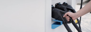 El ele tutuşan EV şarj prizi dışarıda bulanık bir arka plan ile kameranın önünde. Elektrikli araba sürücüsünde şarj prizi vardır. Sürdürülebilir enerji ile çalışan elektrik yükleme istasyonu arka planı