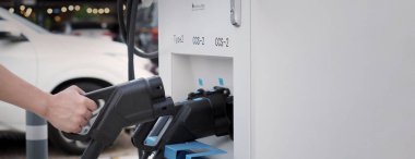 El ele tutuşan EV şarj prizi dışarıda bulanık bir arka plan ile kameranın önünde. Elektrikli araba sürücüsünde şarj prizi vardır. Sürdürülebilir enerji ile çalışan elektrik yükleme istasyonu arka planı