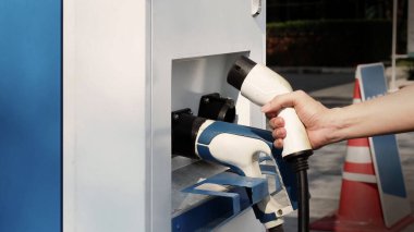 Elektrikli araba sürücüsünde şarj prizi vardır. El ele tutuşan EV şarj prizi dışarıda bulanık bir arka plan ile kameranın önünde. Sürdürülebilir enerji ile çalışan elektrik yükleme istasyonu arka planı