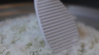 Yakın plan, pilav pişirme makinesinden plastik bir pirinç küreğiyle çıkartılan taze pişmiş beyaz pirinç. Tayland 'ın yumuşak dokusu tanecikleri eritiyor. Ev yapımı pilav pişirme yemekleri. Slow Motion görüntüleri.