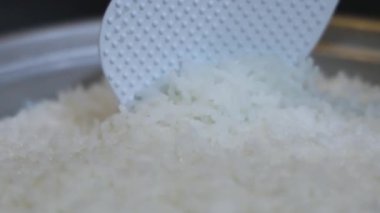 Yakın plan, pilav pişirme makinesinden plastik bir pirinç küreğiyle çıkartılan taze pişmiş beyaz pirinç. Tayland 'ın yumuşak dokusu tanecikleri eritiyor. Ev yapımı pilav pişirme yemekleri. Slow Motion görüntüleri.