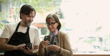 Dost canlısı orta yaşlı bir barista ve kıdemli bir iş kadını modern bir kafede akıllı telefon ve dizüstü bilgisayar kullanarak iş birliği yapıyorlar. Çevrimiçi siparişleri, dijital pazarlama veya iş stratejisini tartışıyor gibi görünüyorlar.