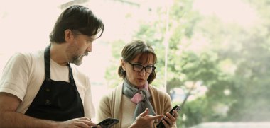 Dost canlısı orta yaşlı bir barista ve kıdemli bir iş kadını modern bir kafede akıllı telefon ve dizüstü bilgisayar kullanarak iş birliği yapıyorlar. Çevrimiçi siparişleri, dijital pazarlama veya iş stratejisini tartışıyor gibi görünüyorlar.