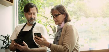 Dost canlısı orta yaşlı bir barista ve kıdemli bir iş kadını modern bir kafede akıllı telefon ve dizüstü bilgisayar kullanarak iş birliği yapıyorlar. Çevrimiçi siparişleri, dijital pazarlama veya iş stratejisini tartışıyor gibi görünüyorlar.