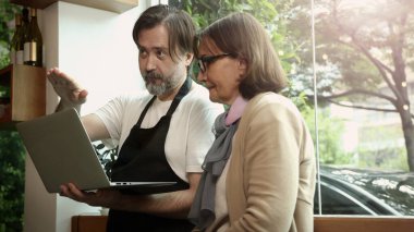 Dost canlısı orta yaşlı bir barista ve kıdemli bir iş kadını modern bir kafede akıllı telefon ve dizüstü bilgisayar kullanarak iş birliği yapıyorlar. Çevrimiçi siparişleri, dijital pazarlama veya iş stratejisini tartışıyor gibi görünüyorlar.