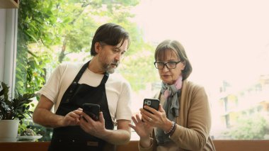 Dost canlısı orta yaşlı bir barista ve kıdemli bir iş kadını modern bir kafede akıllı telefon ve dizüstü bilgisayar kullanarak iş birliği yapıyorlar. Çevrimiçi siparişleri, dijital pazarlama veya iş stratejisini tartışıyor gibi görünüyorlar.