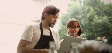 Dost canlısı orta yaşlı bir barista ve kıdemli bir iş kadını modern bir kafede akıllı telefon ve dizüstü bilgisayar kullanarak iş birliği yapıyorlar. Çevrimiçi siparişleri, dijital pazarlama veya iş stratejisini tartışıyor gibi görünüyorlar.
