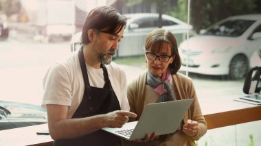 Dost canlısı orta yaşlı bir barista ve kıdemli bir iş kadını modern bir kafede akıllı telefon ve dizüstü bilgisayar kullanarak iş birliği yapıyorlar. Çevrimiçi siparişleri, dijital pazarlama veya iş stratejisini tartışıyor gibi görünüyorlar.