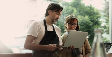 Dost canlısı orta yaşlı bir barista ve kıdemli bir iş kadını modern bir kafede akıllı telefon ve dizüstü bilgisayar kullanarak iş birliği yapıyorlar. Çevrimiçi siparişleri, dijital pazarlama veya iş stratejisini tartışıyor gibi görünüyorlar.
