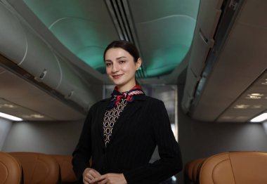 Uçuş görevlisi ya da hostes. Dost canlısı bir uçuş görevlisi, bir uçak kabininin içinde güvenle durur. Profesyonel konukseverlik ve zarif üniforma. Havacılık hizmeti seyahat havayolu hostesi.